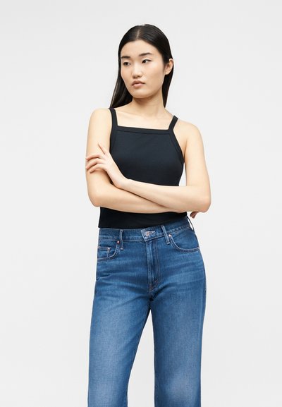 Νεαρή γυναίκα με μακριά σκούρα μαλλιά στέκεται με τα χέρια σταυρωμένα, φορώντας μαύρο αμάνικο τοπ και μπλε ψηλόμεσα τζιν μπροστά σε απλό φόντο.