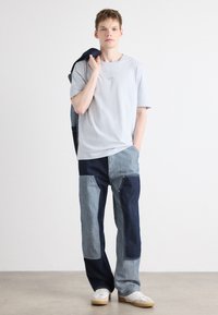Light gray t-shirt, denim patchwork pants in blue tones, featuring varied textures, laid-back fit, and white sneakers with gum soles.