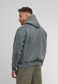 Graue Kapuzenjacke mit einer großen Kapuze, überschnittenen Schultern und gerippten Bündchen. Der Stoff wirkt glatt mit einem lockeren Schnitt und leichtem Glanz.