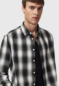 Czarna i biała koszula w kratę z guzikiem, z kołnierzykiem, lekką teksturą i kontrastującymi guzikami. Noszona na ciemnej, okrągło-dekoltowej koszulce.