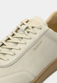 Light beige suede sneaker featuring a textured upper, gold embossed logo, and tan rubber sole with matching laces and stitching.