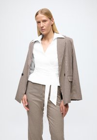 Blazer a quadri marrone e bianco, top avvolgente bianco con nodo, e pantaloni abbinati a quadri. Tessuto liscio e design strutturato con dettagli sartoriali.