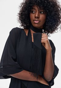 Kobieta o kręconych włosach, ubrana w czarny strój z frędzlami, patrząca w bok, trzymająca szalik przy szyi.