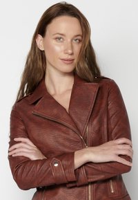 Giacca da biker in pelle marrone con una finitura testurizzata, chiusura con zip asimmetrica e dettagli in hardware argentato. Maniche lunghe e colletto a revers.
