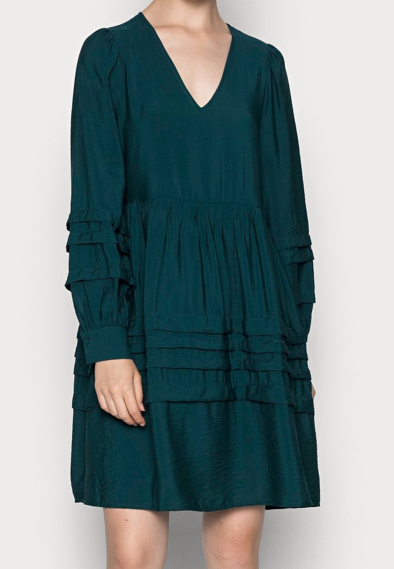 Femme portant une robe longue manches longues vert foncé avec des détails à volants superposés et un décolleté en V profond, visible des épaules aux genoux.