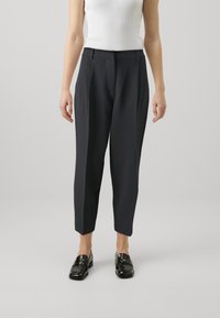 Pantalons fuselés bleu marine avec une texture lisse, présentant une taille mi-haute et des plis à l'avant, associés à des mocassins noirs et un débardeur blanc.