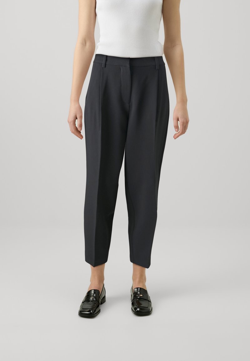 Pantalons fuselés bleu marine avec une texture lisse, présentant une taille mi-haute et des plis à l'avant, associés à des mocassins noirs et un débardeur blanc.