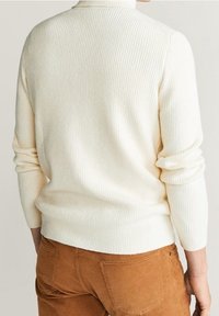 Cremfarbener, gerippter Pullover mit tailliertem Design und hohem Kragen, kombiniert mit braunen Cordhosen. Der Pullover hat lange Ärmel und eine strukturierte Oberfläche.