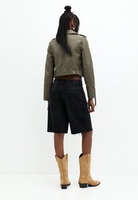 Giacca in pelle verde oliva cropped sopra pantaloni bermuda in denim neri, con orli sfilacciati. Stivali da cowboy color tan con dettagli a contrasto sul tacco e cuciture.