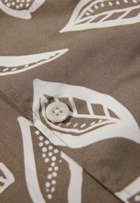 Bouton blanc cousu sur un tissu marron avec de grands motifs de feuilles blanches et brun foncé et des coutures visibles.