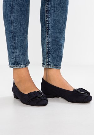 Ballerines en suede marine avec un bout arrondi, un nœud décoratif et un détail de boucle, et un petit talon empilé, associées à un jean bleu clair.