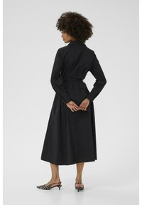 Black long-sleeve dress with a belted waist, buttoned collar, and a flowing skirt, displayed from the back. Worn with striped slingback shoes.