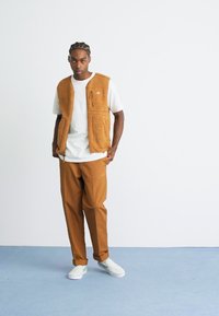 Man with braided hair wearing a white t-shirt, brown fleece vest, brown pants, and white checker slip-on shoes on a blue floor.