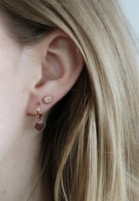 Roségouden oorbellen met een ronde stud en een hangend hartje, beide met een gepolijste afwerking, gedragen aan een geperforeerd oor.