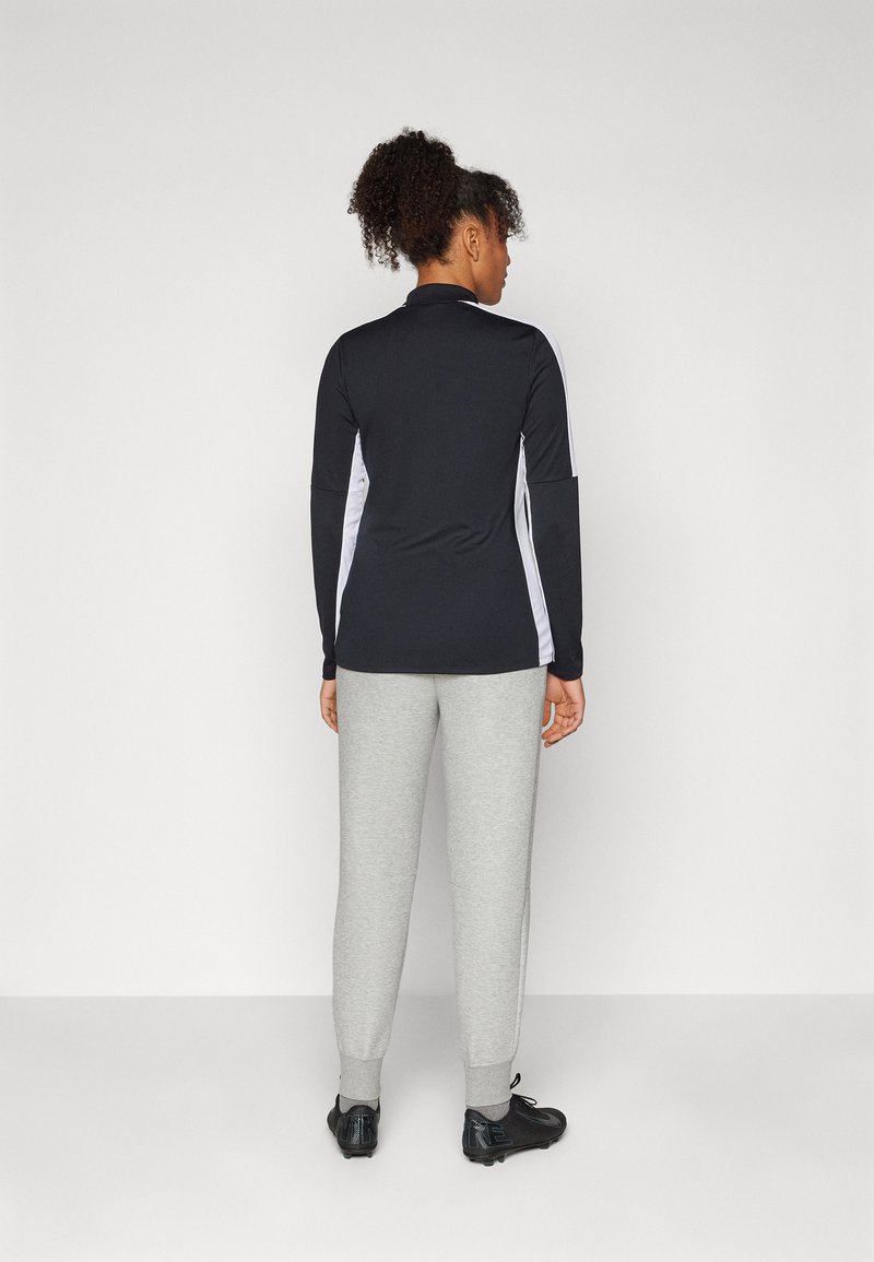 Maglietta sportiva nera a maniche lunghe con pannelli bianchi ai lati, abbinata a pantaloni tuta grigi chiari e scarpe da ginnastica nere, vista da dietro.