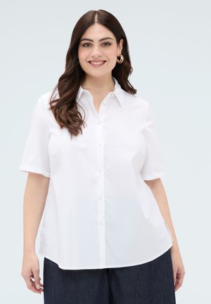 Kobieta o długich brązowych włosach, ubrana w białą koszulę z krótkimi rękawami zapinaną na guziki i ciemne spodnie, uśmiechająca się na jasnym tle.