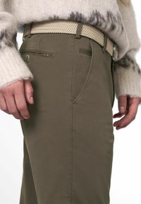 Olijfgroene broek met een gladde textuur, voorzien van voorzakken en een gevlochten beige riem. Geaccentueerd met een metalen knoop nabij de tailleband.