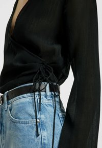 Haut noir texturé à manches longues, avec un col en V et un cordon de serrage à la taille, assorti à un jean en denim bleu clair et une ceinture noire.