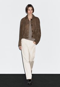 Hnědá semišová bunda s límcem, čtyřmi předními kapsami a černými knoflíky. Nesežená přes texturovaný pletený top a světlé kalhoty.