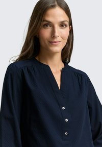 Blusa blu navy realizzata in tessuto testurizzato con scollatura a V, spalle plissettate e quattro piccoli bottoni sul davanti.