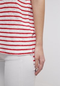 Ruka ženy odpočívající na boku, oblečená v červeném a bílém pruhovaném tričku a bílých kalhotách na jednobarevném pozadí.