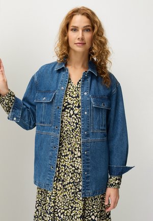 Blaue Jeansjacke mit Fronttaschen und Druckknöpfen, getragen über einem schwarz-gelben gemusterten Kleid mit fließender Silhouette.