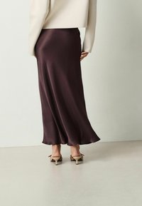 Gonna maxi in satin viola scuro con orlo fluido, abbinata a una maglietta crema e scarpe col tacco taupe, vista da dietro su uno sfondo neutro.