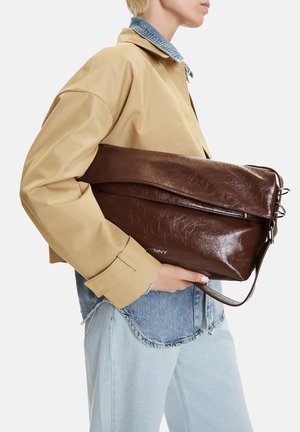 Persona con giacca beige e camicia di jeans che tiene una grande borsa clutch in pelle marrone scuro piegata di lato.