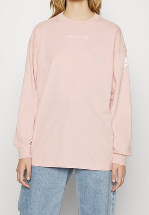 Personne portant une chemise rose clair à manches longues avec le texte "AIR" sur la poitrine et un jean bleu délavé, debout devant un fond blanc.