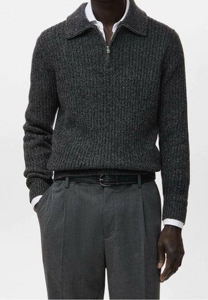 Mann trägt dunkelgrauen, gerippten Reißverschluss-Pullover über weißem Hemd mit Kragen, graue plissierte Hose und schwarzen Gürtel, Hände in den Taschen.