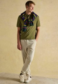 Grünes Polohemd, cremefarbene Cordhosen und weiße Sneaker. Ein bunter Schal mit Streifen in Blau, Grün und Gelb ist um den Hals gelegt.