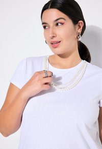 T-shirt bianco a maniche corte indossato con collane di perle a strati. La modella tiene un anello d'argento con dettagli intricati.