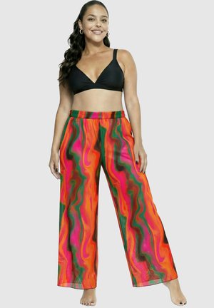 Kobieta uśmiechająca się, ubrana w czarny krótki top i szerokie spodnie z żywym, różowo-pomarańczowo-zielono-czarnym wijącym się wzorem, stojąca boso.