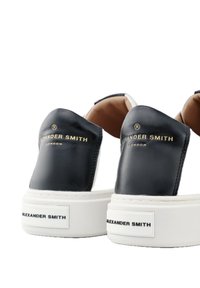 Scarpe slip-on in pelle nera con suola bianca, caratterizzate da un marchio dorato sul tallone e una texture liscia, angolate per la visualizzazione.