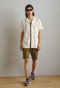 Lyhythihainen nappipaitaja, jossa on valkoisia ja oransseja pystysuoria raitoja, yhdistettynä oliivinvihreisiin shortseihin ja tummaan urheilukenkäpariin.