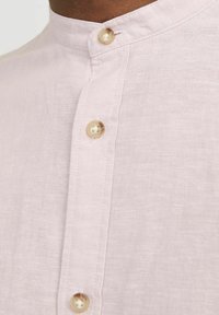 Camicia di lino rosa chiaro con colletto alla coreana, dotata di una chiusura con bottoni e dettagli di bottoni in madreperla sul davanti. Tessuto morbido e con una consistenza piacevole.