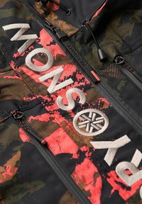 Chaqueta con patrón de camuflaje en verde oscuro, marrón y negro con acentos en rosa brillante. Presenta un tejido texturizado y logotipos en relieve prominentes.