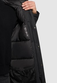 Ruka držící otevřený černý polstrovaný interiér tmavého kabátu, kde je vidět síťová kapsa a štítek s pokyny na údržbu v podšívce.