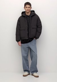 Tânăr stând cu fața la cameră, îmbrăcat într-o geacă puffer neagră, blugi largi de culoare albastru deschis și teniși albi, pe un fundal alb.