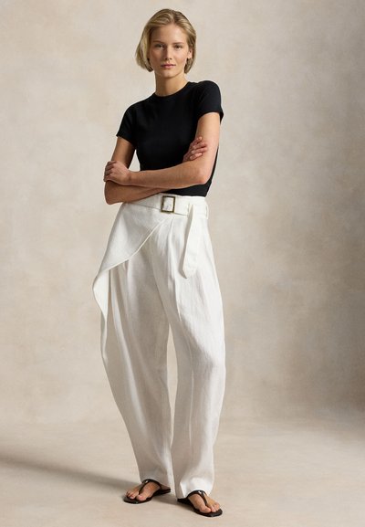 Γυναίκα όρθια με σταυρωμένα τα χέρια, φορώντας μαύρο τοπ με κοντά μανίκια, λευκό παντελόνι με φαρδιά πόδια και ψηλή μέση με ζώνη, και μαύρα σανδάλια.