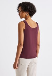Top senza maniche color marrone bordeaux con dettagli arricciati sulle spalle, tessuto morbido e vestibilità comoda, abbinato a pantaloni chiari a vita alta.