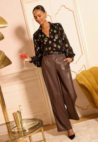 Bluză neagră transparentă cu modele colorate din paiete, pantaloni evazați din piele ecologică maro cu buzunare și un pahar de martini în mână.