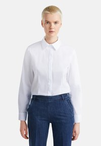 Camicia bianca a maniche lunghe con colletto rigido e texture liscia, abbinata a pantaloni in denim blu con tasche frontali e cuciture.
