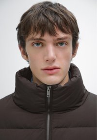 Brązowa pikowana kurtka puchowa z wysokim kołnierzem i zamkiem błyskawicznym z przodu. Charakteryzuje się pikowaną teksturą i eleganckim fasonem. Noszona na ciemnej koszuli.