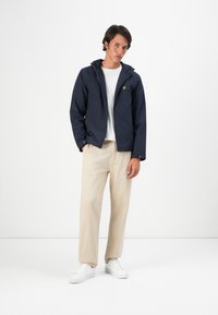 Young man wearing a navy zip-up hooded jacket, white t-shirt, beige pants, and white sneakers standing against a plain white background.