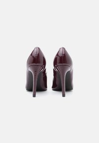 Par de sapatos de salto alto em um tom de borgonha escuro e brilhante vistos de trás sobre um fundo branco.