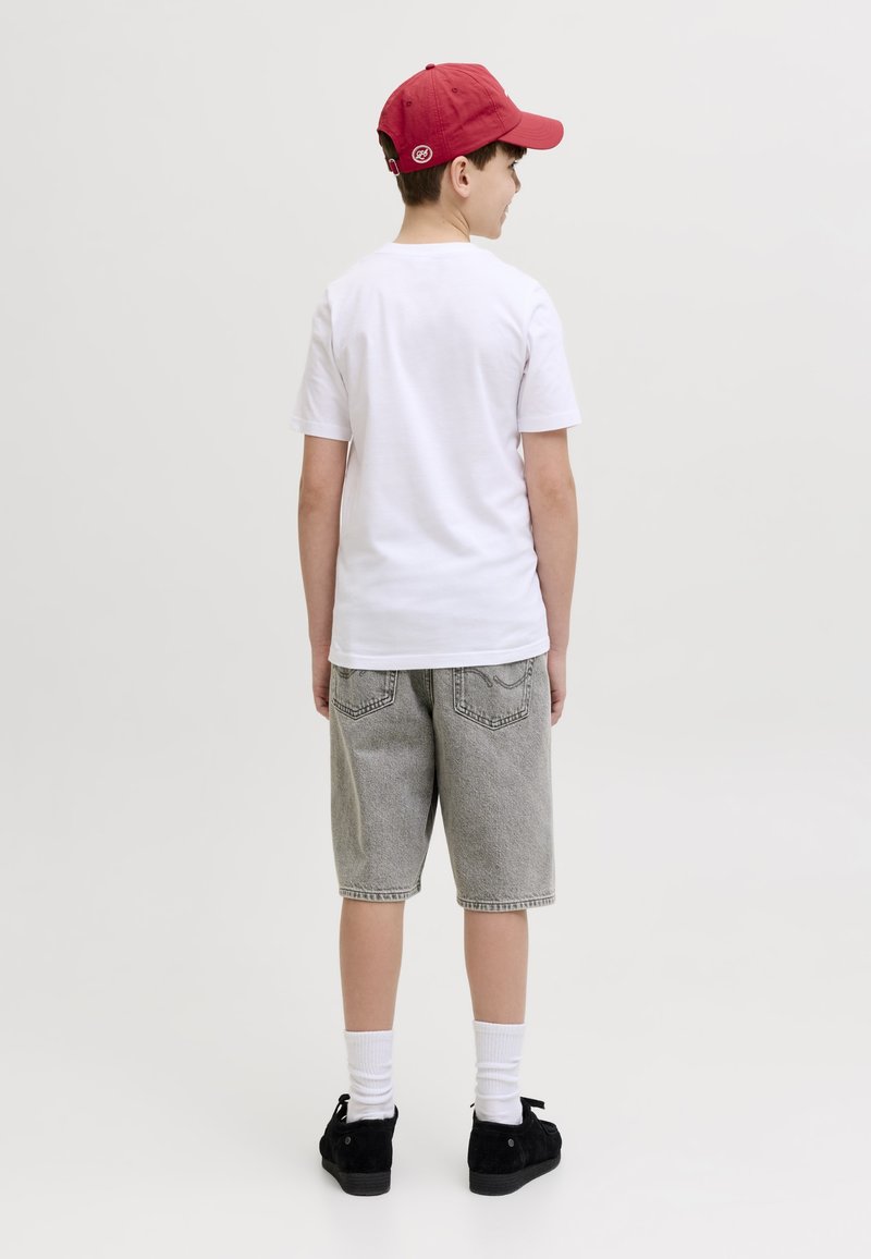 Ragazzo con cappellino rosso, t-shirt bianca, pantaloncini di jeans grigi, calzini bianchi e scarpe nere, in piedi di spalle su uno sfondo semplice.