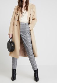 Beige vlněný kabát s velkými knoflíky, spárovaný s šedými kostkovanými kalhotami a černými kotníkovými botami. Oblečení zahrnuje malou černou kabelku.