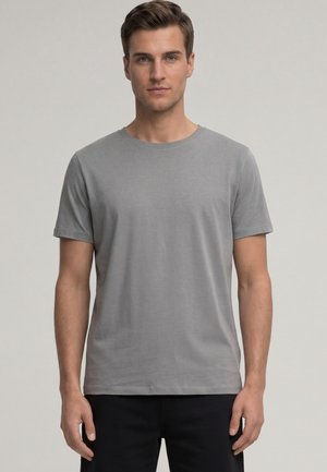 Mann steht frontal, trägt ein schlichtes graues T-Shirt mit kurzen Ärmeln und schwarze Hose vor neutralem Hintergrund.