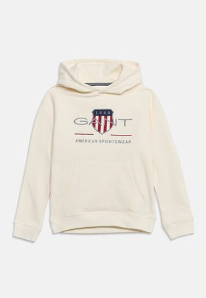 Sweat à capuche crème avec poche kangourou devant, logo GANT, emblème en forme de bouclier avec "1949" et texte "American Sportswear" sur la poitrine.